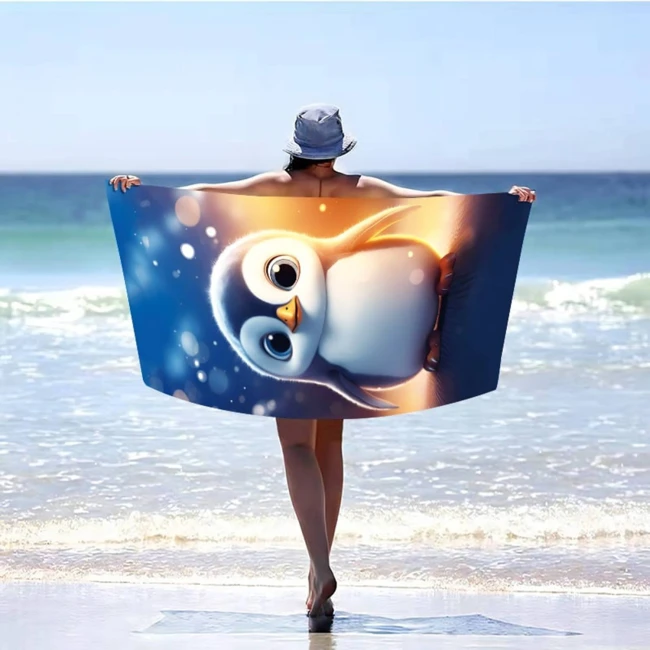 Ręcznik plażowy 100x180 cm z mikrofibry z kolorowym nadrukiem przedstawiającym pingwinka. Produkt trzymany w pozycji pionowej przez osobę w kapeluszu, ustawioną na tle plaży i morza. Morska kolorystyka oraz duży rozmiar idealne do użycia na plaży.