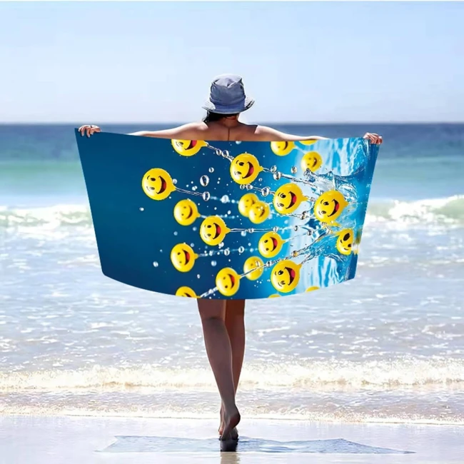 Ręcznik plażowy 100x180 cm w kolorze niebieskim z wzorem żółtych buźek, wykonany z szybkoschnącej mikrofibry. Stojący, rozłożony, trzymany przez osobę na plaży, z wodą w tle.