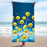 Ręcznik plażowy 100x180 cm w kolorze niebieskim z wzorem żółtych emotikonów z uśmiechami. Produkt wiszący w plecionej formie, na tle morza i plaży, z widocznymi falami. Szybkoschnący materiał mikrofibrowy. Idealny do użycia na plaży lub basenie.
