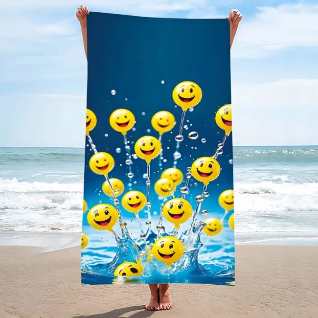 Ręcznik plażowy 100x180 cm w kolorze niebieskim z wzorem żółtych emotikonów z uśmiechami. Produkt wiszący w plecionej formie, na tle morza i plaży, z widocznymi falami. Szybkoschnący materiał mikrofibrowy. Idealny do użycia na plaży lub basenie.
