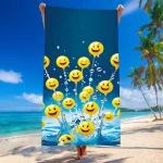 Ręcznik plażowy 100x180 cm w kolorze niebieskim z motywem żółtych buziek. Produkt rozłożony, trzymany w pionie. Widoczny skok wody w tle, palmowe drzewa przy plaży. Materiał mikrofibra, szybkoschnący, idealny na plażę.