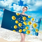 Ręcznik plażowy 100x180 cm wykonany z mikrofibry, z wzorem żółtych buziek na niebieskim tle. Produkt trzymany przez osobę w stroju kąpielowym, leżący na plaży w otoczeniu piasku i morza. Szybkoschnący materiał, duże rozmiary, idealny do użytku na plaży.