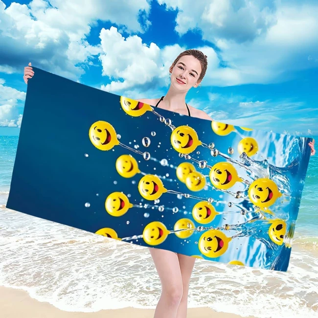 Ręcznik plażowy 100x180 cm wykonany z mikrofibry, z wzorem żółtych buziek na niebieskim tle. Produkt trzymany przez osobę w stroju kąpielowym, leżący na plaży w otoczeniu piasku i morza. Szybkoschnący materiał, duże rozmiary, idealny do użytku na plaży.