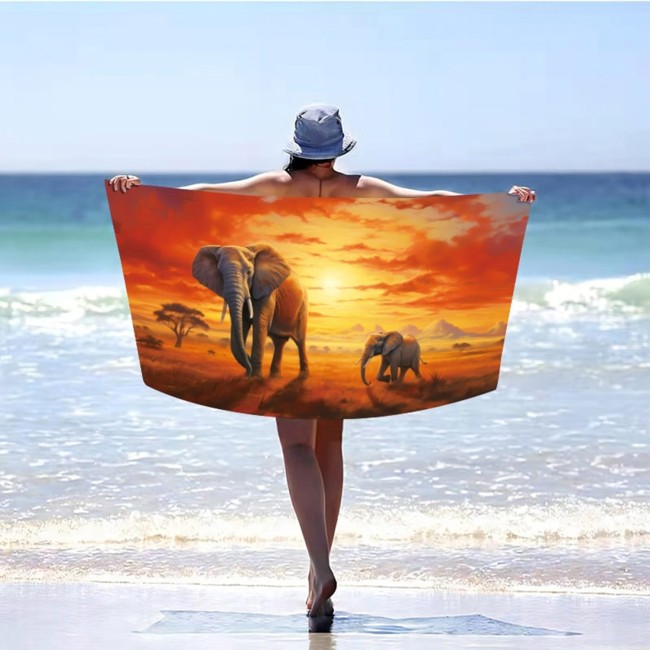 Ręcznik plażowy 100x180 cm z mikrofibry, z grafiką słoni na safari w odcieniach pomarańczowych i żółtych, rozłożony za osobą stojącą na plaży. Produkt szybkoschnący, widoczny w kontekście plażowym, tło z niebieskim morzem i jasnym niebem.