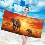 Ręcznik plażowy 100x180 cm w mikrofibrze z grafiką słoni na safari, ukazującą dwa słonie na tle zachodzącego słońca. Produkt trzymany przez osobę w kostiumie kąpielowym na plaży, w otoczeniu piasku i morza. Tekstura gładka, materiał szybkoschnący.