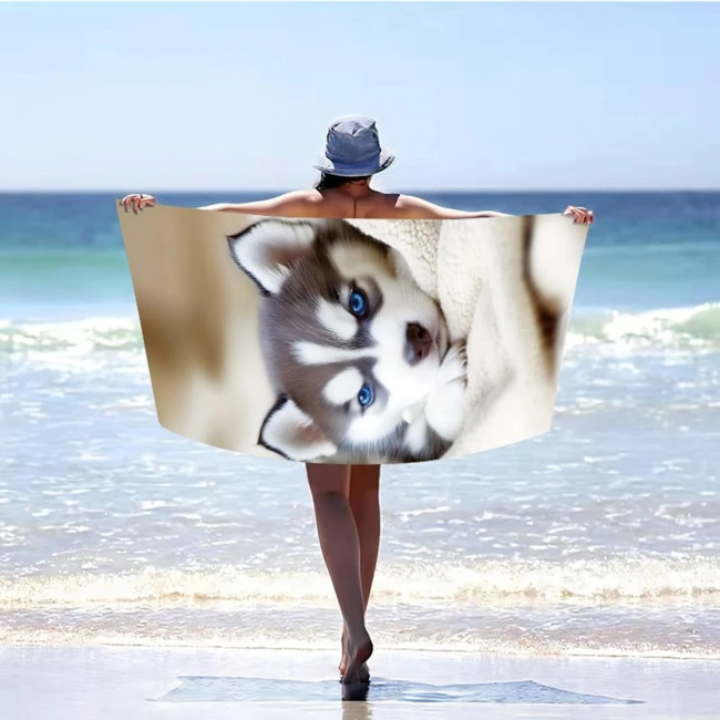 Ręcznik plażowy 100x180 cm z szybkoschnącej mikrofibry, z wizerunkiem szczeniaka husky. Produkt leżący, trzymany przez osobę na plaży, w tle widoczna woda i niebo. Dominujące kolory: brązowy i błękitny.