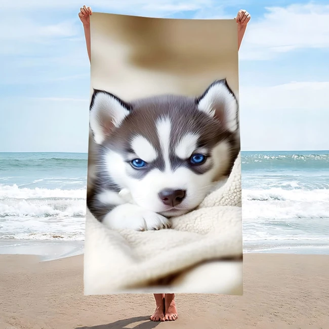 Ręcznik plażowy 100x180 cm z nadrukiem szczeniaka, szybkoschnący, wykonany z mikrofibry. Ujęcie z przodu, produkt trzymany przez osobę na plaży, w tle morze i niebo. Widoczne detale twarzy psa z niebieskimi oczami i białymi akcentami.
