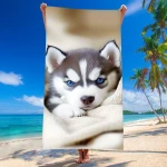 Ręcznik plażowy 100x180 cm z szybkoschnącej mikrofibry, w kolorze beżowym, z nadrukiem szczeniaka husky o niebieskich oczach. Produkt rozłożony, trzymany na plaży, w tle błękitne morze i palmy.
