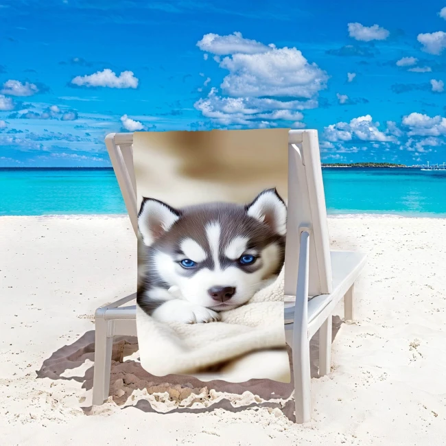 Ręcznik plażowy 100x180 cm z motywem psa husky z błękitnymi oczami, wykonany z mikrofibry. Produkt leży na leżaku w pozycji rozłożonej, z widokiem na plażę i morze w tle, co podkreśla jego letni charakter.