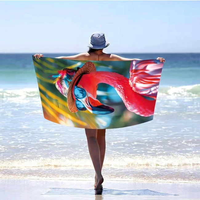 Ręcznik plażowy 100x180 cm z kolorowym wzorem flaminga w okularach, wykonany z mikrofibry. Złożony w dłoniach osoby stojącej na plaży, z widocznym tłem morza i nieba. Szybkoschnący, duży i różowy, idealny na lato.