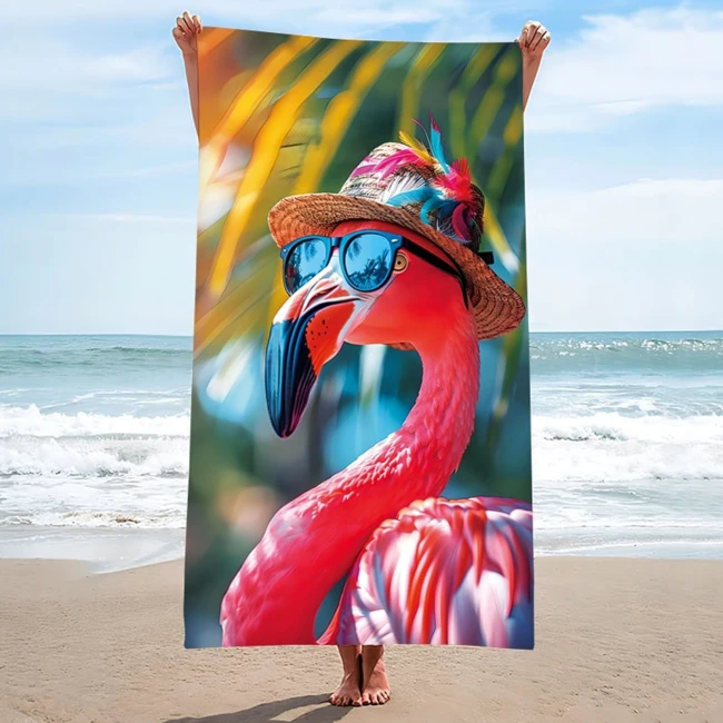 Ręcznik plażowy w kolorze różowym z motywem flaminga w okularach przeciwsłonecznych. Wykonany z szybkoschnącej mikrofibry, o wymiarach 100x180 cm. Ujęcie frontalne, produkt trzymany na plaży, w tle morze i niebo.