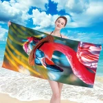 Ręcznik plażowy o wymiarach 100x180 cm z motywem flaminga w okularach, wykonany z szybkoschnącej mikrofibry. Prezentowany w pozycji poziomej, trzymany przez osobę na tle plaży i nieba. Dominujące kolory to różowy i zielony.