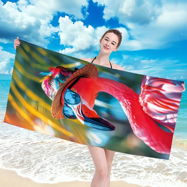 Ręcznik plażowy o wymiarach 100x180 cm z motywem flaminga w okularach, wykonany z szybkoschnącej mikrofibry. Prezentowany w pozycji poziomej, trzymany przez osobę na tle plaży i nieba. Dominujące kolory to różowy i zielony.