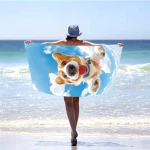 Ręcznik plażowy 100x180 cm z mikrofibry z grafiką psa corgi na błękitnym tle z chmurami, przedstawiony w pełnym fragmencie, trzymany przez osobę stojącą na plaży, w kącie z przodu. Produkt duży, szybkoschnący, idealny na plażę lub basen.