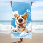 Ręcznik plażowy z mikrofibry 100x180 cm w kolorze błękitnym, z wizerunkiem psa rasy corgi na tle nieba i chmur. Produkt trzymany jest w pozycji pionowej przez osobę stojącą na plaży, z morzem w tle. Idealny do użytku na plaży.