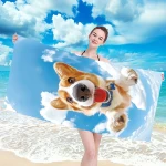 Ręcznik plażowy 100x180 z mikrofibry, z grafiką psa na tle błękitnego nieba i chmur. Produkt leży rozłożony w dłoniach kobiety na tle plaży, co podkreśla jego zastosowanie. Materiał szybkoschnący.