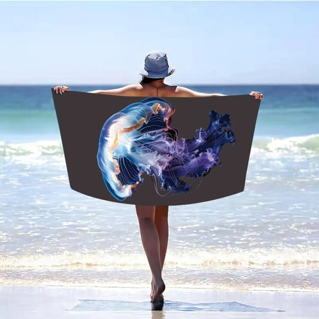 Ręcznik plażowy 100x180 cm, wykonany z mikrofibry. Na tkaninie widoczny motyw meduzy w odcieniach błękitu i fioletu. Produkt leży w rękach osoby stojącej na plaży, w tle morze i jasne niebo. Ręcznik jest rozłożony, ukazując swój pełny wzór.