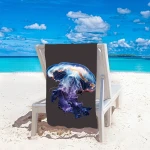 Ręcznik plażowy 100x180 cm z mikrofibry, z grafiką meduzy w odcieniach niebieskiego i fioletowego, położony na leżaku na plaży, w tle turkusowa woda i jasny piasek, ukazujący detale wzoru i teksturę materiału.