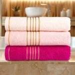 Ręcznik kąpielowy bawełniany 50x90 cm w kolorze różowym, z bordiurą w złote paski. Układ poziomy, leżący na kamiennej powierzchni. Widoczny jeden różowy i dwa różne odcienie jasnoróżowego, tekstura puszysta. Tło w łazience z oświetleniem.