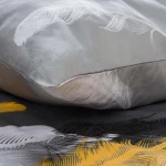 Pościel satyna bawełniana 140x200 cm w kolorze szarym z białymi i złotymi piórami, leży nałożona na podkład szary. Wyróżniająca się delikatna, gładka faktura tkaniny, podkreślająca elegancki wzór piór. Fragment poduszki w zbliżeniu.