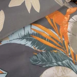 Pościel satyna bawełniana 180x200 w tropikalne liście na szarym tle. Produkt leży w złożeniu, ukazując kolorowe wzory liści w odcieniach zieleni, pomarańczu i beżu. Materiał jest gładki, a tło ma jednolitą szarość, co podkreśla więzów wzorzystych.