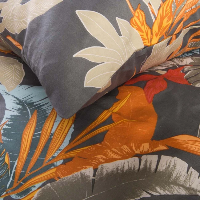 Pościel satyna bawełniana 220x200 Tropenza w kolorze szaro-beżowym z tropikalnymi liśćmi. Widoczne poduszki i narzuta z różnorodnymi wzorami liści w odcieniach zieleni, pomarańczu i czerwieni.