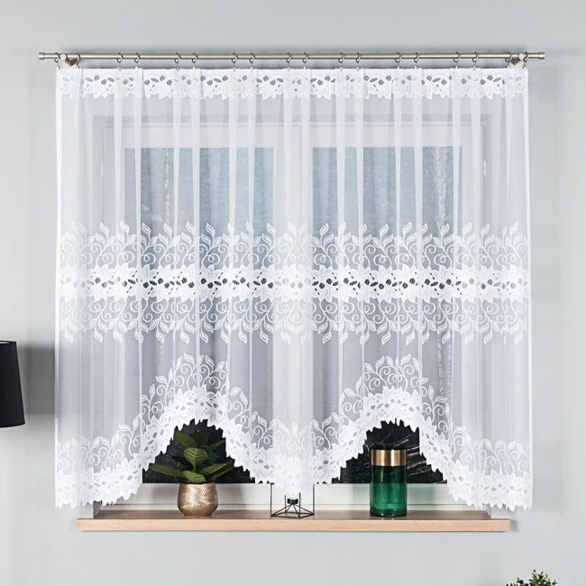 Firana gotowa BASTIA żakardowa, 300x160 cm, w kolorze białym, wisząca na karniszu. Wzór z kwiatowymi detalami na dolnej krawędzi oraz przejrzysta tkanina nadają lekkości. Produkt umieszczony na tle jasnej ściany, z elementami dekoracyjnymi na parapecie.