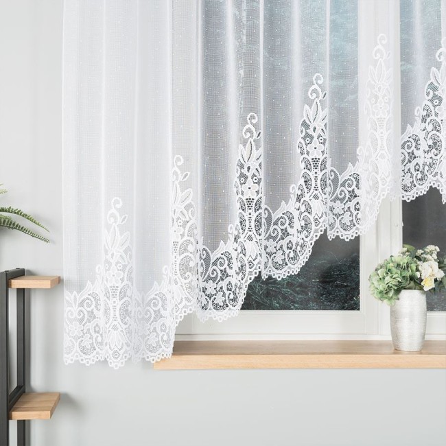 Firana gotowa BERENIKA żakardowa, o wymiarach 400x160 cm, w kolorze białym. Zawieszona na oknie, z haftowanym wzorem na dolnej krawędzi. Tło neutralne, z wazonem oraz rośliną w pobliżu. Przez firanę prześwituje światło, nadając przytulności wnętrzu.