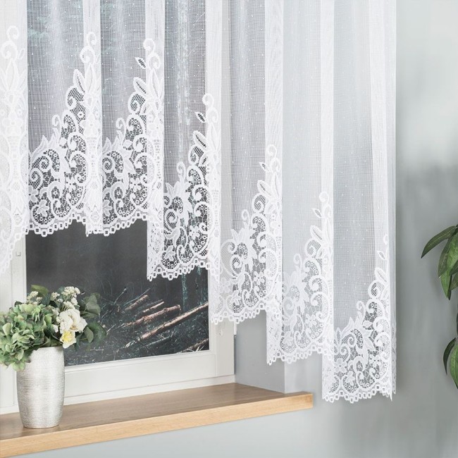 Firana gotowa BERENIKA w kolorze białym, wykonana z żakardowego materiału, o wymiarach 400x160 cm. Zawieszona na oknie, z dekoracyjnym, ażurowym wzorem na dolnej krawędzi. Tło z drewnianą półką i doniczką z rośliną.