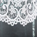 Firana gotowa BERENIKA żakardowa, w kolorze białym, z subtelnym wzorem. Zbliżenie na dolną krawędź, ukazującą koronkowe zdobienia. Materiał lekki, teksturowany, idealny do okna. Tło w odcieniach szarości, minimalistyczne. Wymiary 400x160 cm.