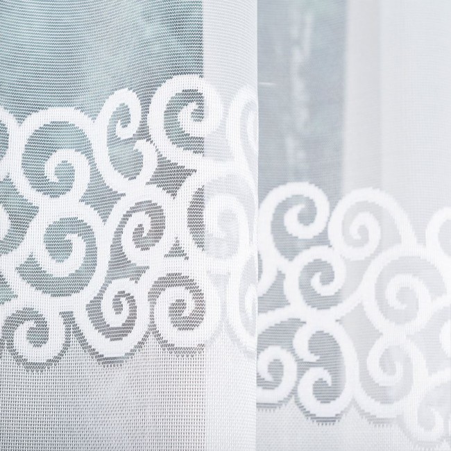 Biała firana gotowa DANIELA z żakardowym wzorem, o szerokości 300 cm i wysokości 140 cm, wisząca na oknie. Wzór z falistymi spiralami jest wpleciony w tkaninę, podkreślający jej teksturę i przezroczystość. Tło minimalistyczne.