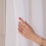 Firana gotowa ETAMINA w kolorze białym na srebrnych przelotkach. Zbliżenie na tkaninę, widoczne detale tekstury. Produkt zawieszony, ręka z pierścionkiem dotyka firany. Tło jasne, jednolite. Rozmiar 200x250 cm.