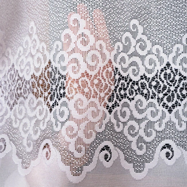 Firana żakardowa MODESTA w kolorze białym, o wymiarach 200x250 cm. Widoczna finezyjna siateczka z wzorem w formie spiralnych zdobień oraz falistym wykończeniem. Produkt rozłożony, zbliżenie na detal faktury tkaniny.