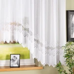 Firana gotowa MODESTA, żakardowa, biała, o wymiarach 500x160 cm, zawieszona na karniszu, z dekoracyjnym brzegiem oraz delikatnym wzorem. Tło gładkie, z widocznym drewnianym parapetem i ramką na zdjęcie. Wnętrze jasne, z roślinnością w tle.