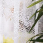 Firana gotowa MODESTA żakardowa, w kolorze białym, o wymiarach 500x160 cm. Zbliżenie na teksturę tkaniny z ornamentalnym wzorem. Produkt wiszący na karniszu, częściowo zasłaniający okno, w tle widoczne zielone liście roślin.