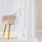 Firana gotowa NALA w kolorze białym, o wymiarach 140x250 cm, wisząca na przelotkach. Materiał lekki, półprzezroczysty, z zasłoną ukazującą załamanie tkaniny. W tle widoczny jasny fotel z miękkim siedziskiem i złotymi nogami, na jaśminowym dywanie.