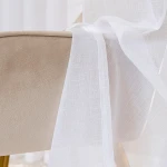 Firana gotowa NALA w kolorze białym, wykonana z lekkiego, teksturowanego materiału, rozpościera się na krawędzi mebla obitego tkaniną. Widoczny detal przelotek ułatwiających zawieszenie. Ujęcie zbliżone, ukazujące delikatność i transparentność firany.