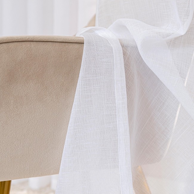 Firana gotowa NALA w kolorze białym, wykonana z lekkiego, teksturowanego materiału, rozpościera się na krawędzi mebla obitego tkaniną. Widoczny detal przelotek ułatwiających zawieszenie. Ujęcie zbliżone, ukazujące delikatność i transparentność firany.