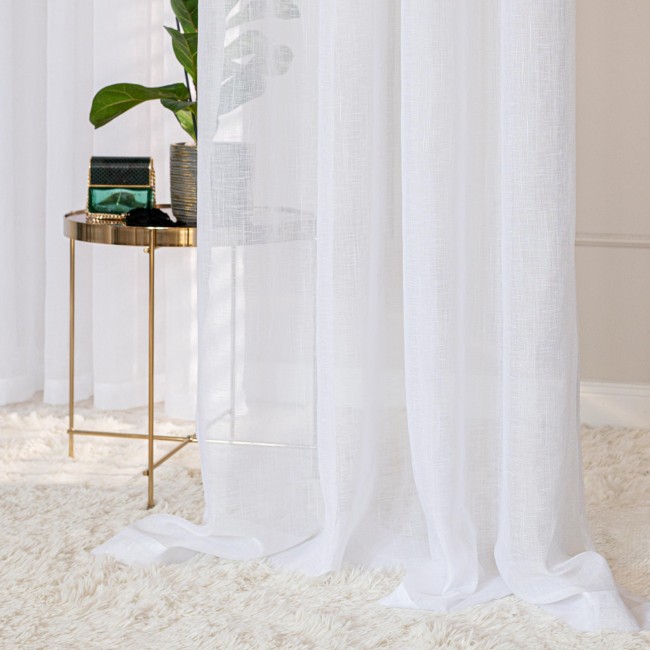 Firana gotowa NALA w kolorze białym, rozmiar 400x250 cm, wykonana z lekkiego materiału, wisząca na przelotkach. Widoczne w tle metalowe stoisko z rośliną oraz dekoracyjnym pudełkiem. Tło utrzymane w stonowanej kolorystyce.