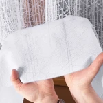 Firana gotowa WIKTORIA żakardowa, biała, o wymiarach 200x250 cm, pokazana w zbliżeniu. Tkanina z delikatnym wzorem żakardowym, trzymana rękami. Produkt w białym kolorze, zaprezentowany na prosto, ukazuje lekkość materiału i teksturę.