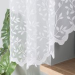 Firana gotowa ZUZANNA w kolorze białym, żakardowa z całopowierzchniowym wzorem liściastym. Ujęcie z boku, wisząca, zakończona koronkowym wykończeniem. Produkt umieszczony na tle szarego muru, obok zielonej rośliny doniczkowej. Wymiary: 300x160 cm.
