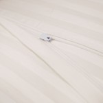 Pościel satyna bawełniana w kolorze kremowym, z widocznym zamkiem błyskawicznym. Ujęcie w zbliżeniu, skupiające się na detalu mocańca, na tle jasnego materiału o delikatnych, poziomych prążkach. Produkt leży płasko.