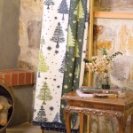 Dwustronny koc bawełniany 150x200 cm w kolorze zielonym, pokryty wzorem choinek i śnieżynek, z frędzlami na brzegach. Produkt leży złożony na drewnianym stoliku, obok znajduje się wazon z suszonymi roślinami oraz książkami.