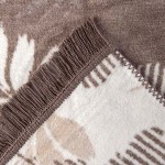 Dwustronny koc bawełniany 150x200, brązowy z wzorem liści i frędzlami. Ujęcie z bliska na fragment koca, ukazujące teksturę i detale wykończenia. Koc leży na gładkim tle, podkreślając jego wzory i fakturę.