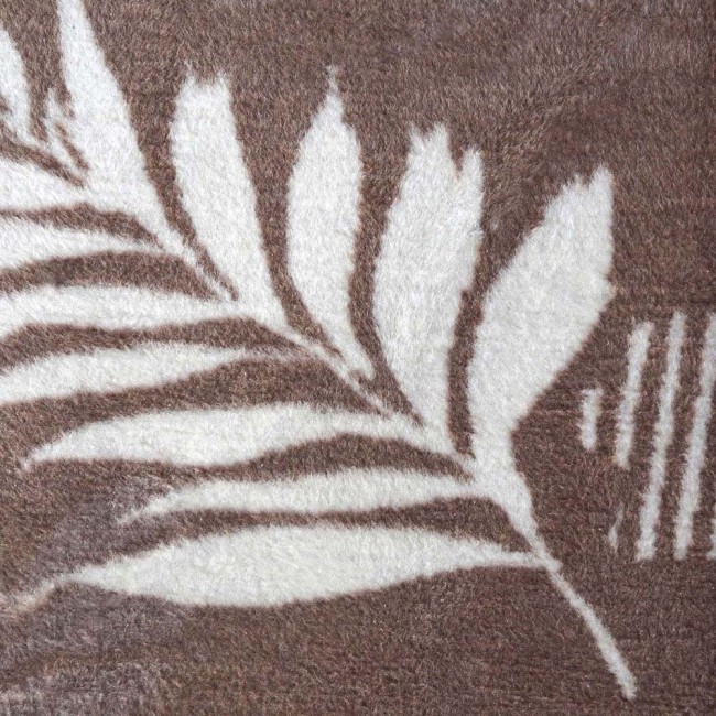 Koc bawełniany 150x200 cm w brązowym kolorze z białym wzorem liści. Dwustronny materiał z frędzlami, leżący na płasko. Wzór roślinny dodaje naturalnego charakteru, a faktura jest miękka i przyjemna w dotyku.