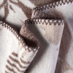 Koc bawełniany 150x200 w brązowe liście, dwustronny. Zwinięty w kształt fali, z widocznymi frędzlami na brzegach. Tekstura miękka, z asymetrycznym wzorem. Ujęcie z bliska ukazujące detale szycia.