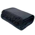 Koc bawełniany 150x200, ciemnoszary, zwinięty z frędzlami na jednym końcu. Ujęcie z boku, eksponujące teksturę materiału. Prosty, jasny podkład, który podkreśla kolor i detale frędzli. Idealny do użytku w domu lub na tarasie.