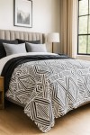 Narzuta drukowana Tarik 170x210, w biało-czarnym wzorze geometrycznym, leży na łóżku. Widoczny kąt ujęcia z boku eksponuje fakturę materiału. Obok narzuty znajdują się szare poduszki, a tło to jasne wnętrze sypialni z oknem i lampą.