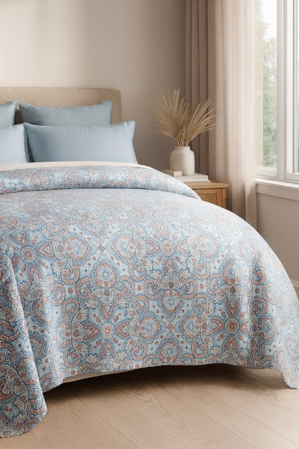 Narzuta drukowana Harem 170x210 w odcieniach błękitu z ornamentalnym wzorem w kolorze miedzi i beżu, rozłożona na łóżku. Kąt ujęcia frontalny, ukazujący detale faktury. W tle widoczne poduszki w błękitnym kolorze oraz drewniana komoda.