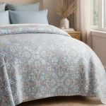 Poszewka na poduszkę 40x40 w kolorze błękitnym z nadrukiem w kwiatowe wzory, zbliżenie na detale tkaniny. Produkt leży na łóżku, obok widoczne dwa poduszki w jasnym kolorze. Tło neutralne, w przytulnej aranżacji sypialni.